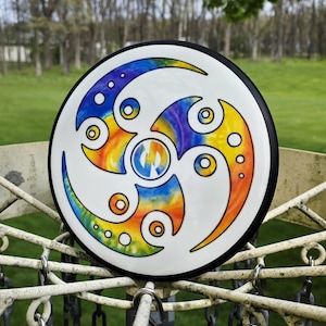 May include: A white disc golf disc with a colorful, abstract design. The design features a swirling pattern in shades of blue, orange, yellow, and purple, with a black rim. The disc is set against a backdrop of green grass and trees.
