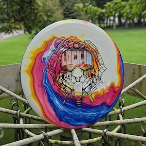 Pode incluir: Um disco de golfe branco com um padrão tie-dye vibrante e rodopiante em tons de rosa, amarelo e azul. O gráfico central apresenta uma ilustração detalhada a preto e branco de uma figura com capacete e o texto LOCK IN na viseira.