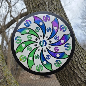 May include: A colorful disc golf disc with a black rim. The disc features a white center with a spiral design of green, blue, and purple shapes, each containing the letter "S". The disc is hanging on a tree.