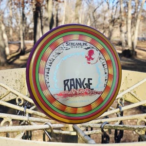 Puede incluir: Un disco de golf de colores con un borde morado y un diseño con la palabra "RANGE" en negrita. El disco tiene un patrón de rayas multicolores y un gráfico de una figura roja. También es visible la marca "STREAMLINE DISCS".