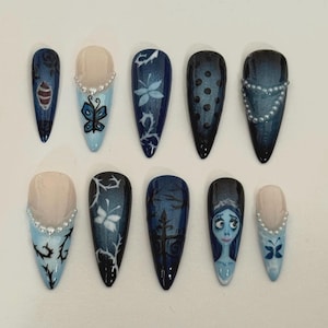 Op de afbeelding: Een set van tien kunstnagels met een donkerblauwe en lichtblauwe kleurstelling. Elke nagel heeft unieke ontwerpen, waaronder bloempatronen, stippen en een portret van een personage. Sommige nagels zijn versierd met kleine, parelachtige versieringen.