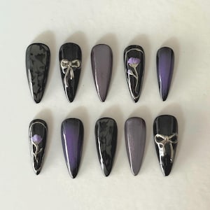Puede incluir: Un conjunto de diez uñas artificiales con varios diseños. Algunas son negras lisas, otras tienen un degradado de morado y negro. Dos uñas presentan lazos plateados y dos diseños florales plateados.