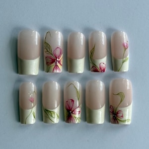 Pode incluir: Um conjunto de dez unhas artificiais com base rosa claro e ponta verde pálido. Cada unha apresenta um desenho floral pintado à mão com flores rosa e amarelas e folhas verdes.
