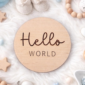 Peut inclure: Une pancarte ronde en bois avec l'inscription "Hello WORLD" en écriture cursive. L'image présente des articles pour bébés, dont des chaussons en tricot, une étoile en bois, un anneau de dentition lapin et une attache-tétine.