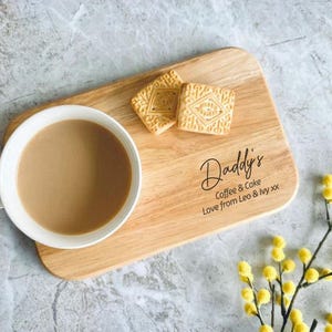 May include: A wooden serving tray with a white mug of coffee and two biscuits. The tray has the text "Daddy's Coffee & Cake Love from Leo & Ivy xx". Yellow flowers are in the bottom right corner.