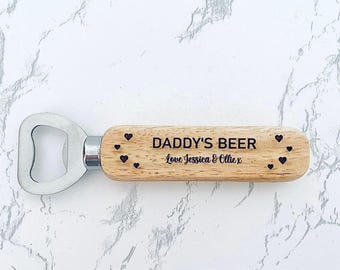 Puede incluir: Un abridor de botellas de madera con cabezal de acero inoxidable. El mango está grabado con las palabras "DADDY'S BEER" y "Love Jessica & Ollie x", junto con varios diseños de pequeños corazones. El abridor está sobre un fondo de mármol blanco.