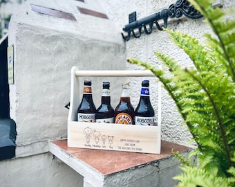 May include: A wooden beer caddy with four bottles of beer. The caddy has a handle and is decorated with a family illustration and text that reads "Happy Father's Day, Love From The Reasons You Drink!"
