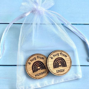 Op de afbeelding: Twee houten tokens met een regenboogontwerp en de woorden "A hug from MUMMY" en "A hug from EMILY" in een witte mesh zak met een trekkoord. De tokens zijn rond en hebben een natuurlijke houtnerf.