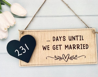 以下が含まれることがあります： 「... DAYS UNTIL WE GET MARRIED」の文字とハートのデザインが施された木製サイン。黒いハート型の黒板には数字の「231」が表示されています。サインは麻ひもで吊り下げられています。