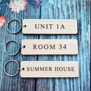 以下が含まれることがあります： 銀色のリングが付いた3つの木製キーホルダー。「UNIT 1A」、「ROOM 34」、「SUMMER HOUSE」の文字が刻印されています。キーホルダーは長方形で、明るい色の木材で作られており、青い木製の背景に配置されています。