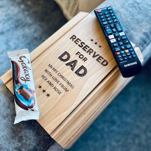 Pode incluir: Uma bandeja de madeira com o texto "RESERVED FOR DAD" e "MERRY CHRISTMAS WITH LOVE FROM REX AND ROSE". Uma barra de chocolate Galaxy e um controle remoto Samsung estão na bandeja. A bandeja está em uma superfície cinza.