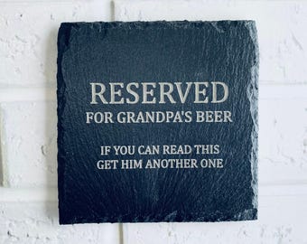 May include: A square, dark gray slate coaster with the text "RESERVED FOR GRANDPA'S BEER" and "IF YOU CAN READ THIS GET HIM ANOTHER ONE" engraved in white. The coaster has a textured surface and is likely for home use.