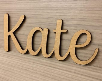 May include: Wooden sign spelling the name "Kate" in a cursive font. The letters are a light brown color with a darker brown outline, mounted on a light wood-grain background. The sign is a decorative item.
