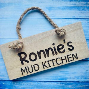 May include: A rectangular wooden sign with the text "Ronnie's MUD KITCHEN" in black lettering. The sign has a natural wood grain finish and a knotted rope handle. The background is a blue wooden surface.