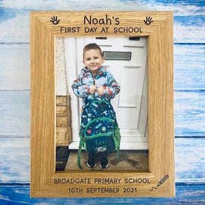 May include: A wooden picture frame with the text "Noah's FIRST DAY AT SCHOOL" and "BROADGATE PRIMARY SCHOOL 10TH SEPTEMBER 2021". The frame holds a photo of a child with a backpack.
