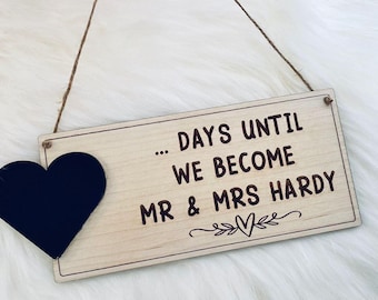 May include: A wooden sign with the text "... DAYS UNTIL WE BECOME MR & MRS HARDY" and a black heart. The sign is rectangular with a light wood grain finish and a decorative border. A twine string is attached for hanging.