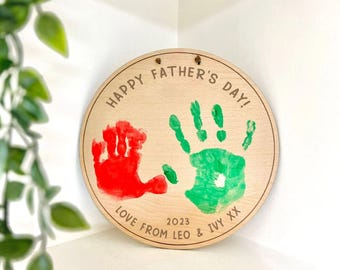 May include: A round wooden plaque with the text "HAPPY FATHER'S DAY!" and handprints in red and green. The text "LOVE FROM LEO & IVY XX 2023" is also included. The plaque has two small holes at the top for hanging.