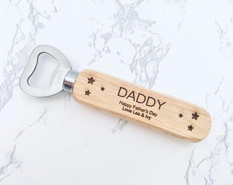 May include: A wooden bottle opener with a stainless steel head. The handle is engraved with the word "DADDY", the message "Happy Father's Day Love Leo & Ivy", and star designs. The opener is on a white marble surface.