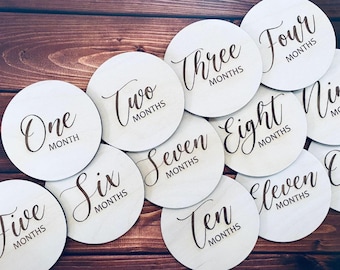 Può includere: Un set di dischi in legno per segnare le tappe mensili di un bambino. Ogni disco rotondo presenta il numero del mese, come "Un mese" e "Due mesi", scritto in un elegante carattere. I dischi sono disposti su una superficie di legno scuro.