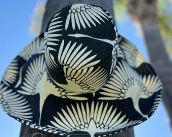 May include: A black and white sun hat with a wide brim and a patterned design of stylized birds. The hat is made of fabric and has a sewn seam around the brim.