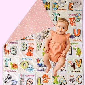 May include: A baby girl is lying on a white and pink alphabet playmat with colorful letters and animal illustrations. The mat features the alphabet from A to Z with corresponding animal illustrations. The mat is made of soft, comfortable fabric and is perfect for babies and toddlers.