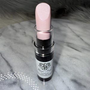 May include: A pink lipstick in a black tube with a clear top. The tube has a white label with the text "Blanc Perle Lipstick" and "Luxhian Beauty" on it.