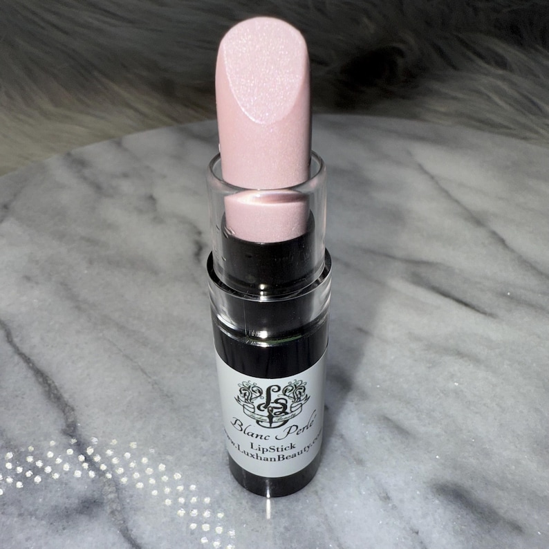 May include: A pink lipstick in a black tube with a clear top. The tube has a white label with the text "Blanc Perle Lipstick" and "Luxhian Beauty" on it.