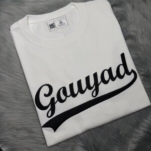 Puede incluir: Una camiseta blanca de cuello redondo con la palabra "Gouyad" en una fuente negra. La palabra está subrayada con una línea negra brillante. La camiseta tiene una etiqueta en el cuello que dice "MAKLINE".
