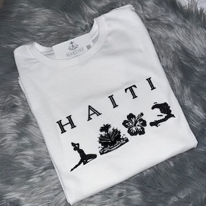 May include: White t-shirt with black text that reads "HAITI" and features a silhouette of a person, a palm tree, a hibiscus flower, and the outline of the island of Hispaniola.
