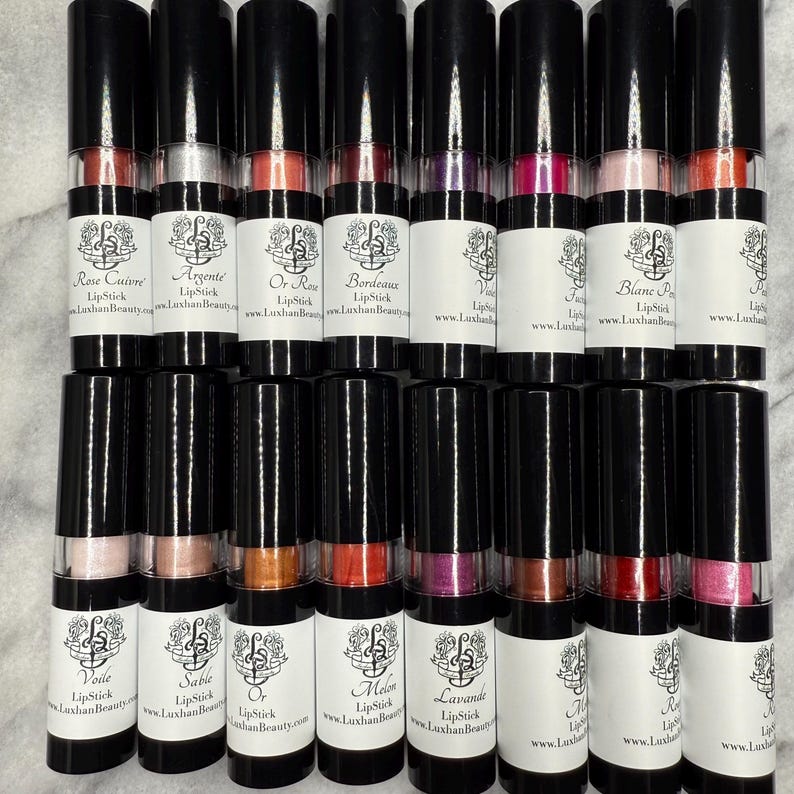 May include: A collection of twenty lipstick tubes, each containing a different shade of lipstick. The tubes are black with a clear window to show the colour of the lipstick. Each tube has a white label with the name of the lipstick colour and the website address "www.LuxhanBeauty.com".