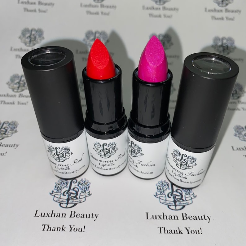 May include: Three lipstick tubes with black caps and white labels. The lipsticks are in the colors red, fuchsia, and a darker pink. The labels read "Shimmering Red Lipstick" and "Shimmering Fuchsia Lipstick". The text "Luxhan Beauty Thank You!" is printed on the white surface below the lipsticks.