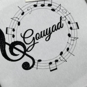 May include: White fabric with black embroidered musical notes and a treble clef. The word "Gouyad" is embroidered in a cursive font within a circle of musical notes. The design is centered on the fabric.