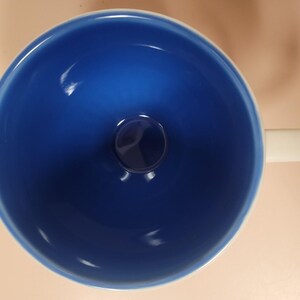 May include: A top-down view of a ceramic mug with a deep blue interior and a white handle. The mug's interior has a glossy finish, and the handle is rectangular. The mug sits on a light tan surface.