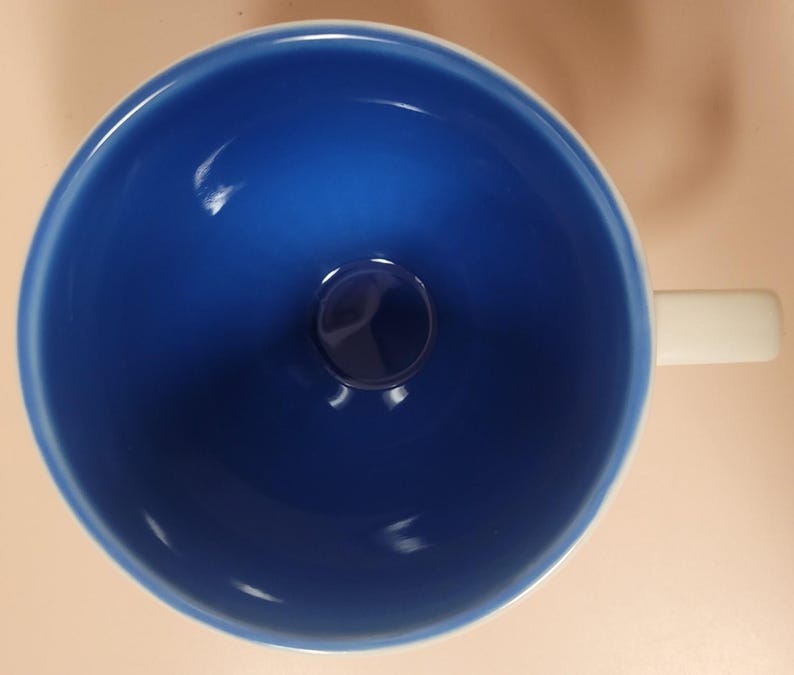 May include: A top-down view of a ceramic mug with a deep blue interior and a white handle. The mug's interior has a glossy finish, and the handle is rectangular. The mug sits on a light tan surface.