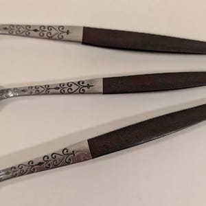 May include: Close-up of three spoons. The spoons have a silver-colored bowl and handle with a dark brown handle. The silver handle has a decorative scroll design. The spoons are arranged diagonally.