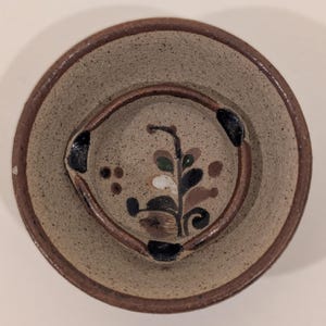 May include: Handmade ceramic plate with a raised center and a wide rim. The plate features a floral design in shades of brown, green, and white. The rim and center ring are a darker brown. The plate is a neutral beige color.