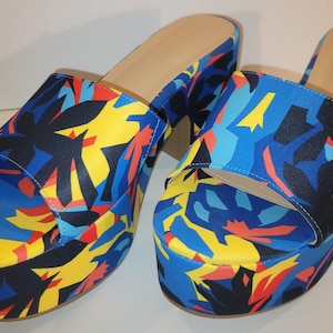 May include: A pair of platform sandals with a colorful floral pattern. The sandals feature a blue, yellow, red, and black design on the straps and platform. The soles are a neutral beige color.