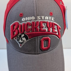 May include: A gray and red baseball cap featuring the embroidered text OHIO STATE BUCKEYES in red and white, a small mascot graphic, and a red number 0 on the front. The cap has a structured crown, a curved brim, and a small white tag on the side.