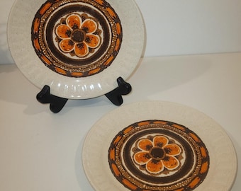 May include: Two off-white ceramic plates with a brown and orange floral design. The plates have a speckled texture and a raised rim. One plate is displayed on a black plate stand. The floral design is centered on the plate.