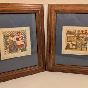 May include: Two framed prints with a blue matting and brown wooden frames. The prints depict quaint, colorful scenes of family life. One shows a family with a man playing a stringed instrument. The other shows a child in a rocking chair.