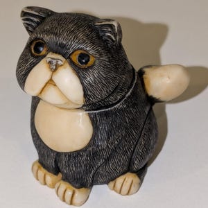 May include: A small, black and white ceramic figurine of a cat with a wide, flat face and large, round eyes. The cat is sitting with its tail curled around its body.