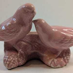 May include: A ceramic figurine featuring two pink birds perched on a textured branch base. The birds have a glossy finish with a visible crackle glaze pattern throughout the surface. The sculpture depicts the birds facing each other closely.