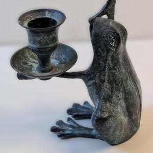 May include: A bronze-colored frog-shaped candle holder. The frog is in a seated position, holding a candle cup and plate. The candle holder has a weathered, antique appearance, with green and blue hues.