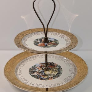 May include: Two-tiered serving tray with a dark brown metal handle. The plates have a gold rim and a central image of people in a garden. The plates are white with a gold filigree design.