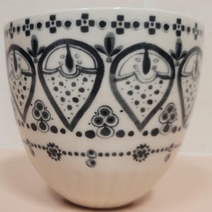 May include: White ceramic cup with a black and gray floral pattern. The design features stylized teardrop shapes filled with dots and outlined with black lines. The cup has a rounded shape and a smooth, glossy finish.