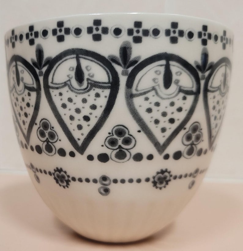 May include: White ceramic cup with a black and gray floral pattern. The design features stylized teardrop shapes filled with dots and outlined with black lines. The cup has a rounded shape and a smooth, glossy finish.