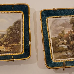 May include: Two square framed decorative plates with gold trim and a teal blue background. Each plate features a different landscape scene with a white background.
