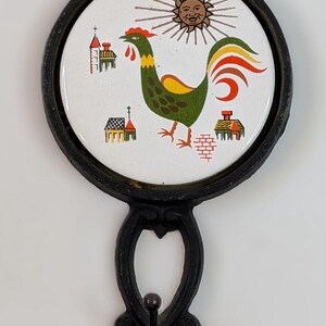 May include: Black cast iron wall hanging with a decorative rooster design. The circular ceramic center features a colorful rooster, sun, and buildings. The piece includes a hook for hanging and a fork-like design.