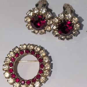 May include: A vintage jewelry set featuring a pair of clip-on earrings and a brooch. The earrings and brooch have a floral design with a large, round, ruby-red center stone surrounded by clear, sparkling crystals. The brooch is a circular design with a gold-tone pin.