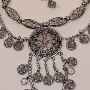 May include: A silver-toned necklace set featuring intricate floral and geometric designs. The set includes a choker, a longer necklace with a large central pendant, and a delicate chain with clear crystal accents. The necklaces have a vintage or bohemian aesthetic.